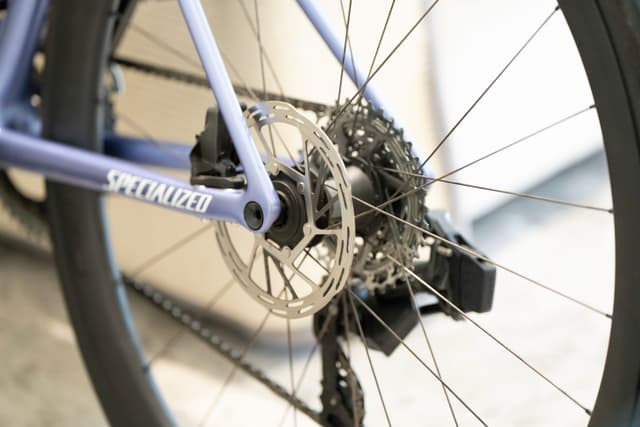 自行车后轮的特写，重点是碟刹、齿轮组和后拨链器。车架是淡紫色，轮辐清晰可见。