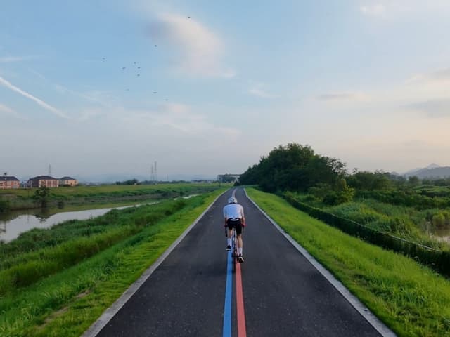 一名背影骑车人沿着带红蓝中线的乡间骑行道前行，两侧绿草堤坝与水道相伴，远处树木与淡淡晚霞的天空相接。