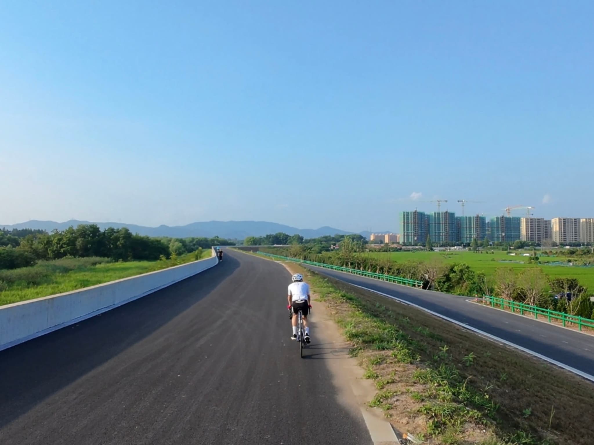 一名骑行者沿着宽阔的柏油路向前骑行，路两侧是绿地和田野，远处有几栋高层建筑，天空晴朗湛蓝。